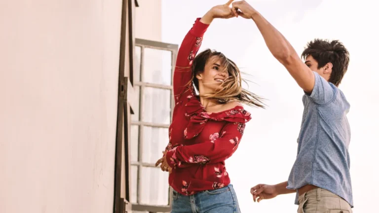 Fotografie ukazuje pár tančící před zdí s otevřeným oknem. Žena v červené květinové halence a džínách zvedá ruku a usmívá se, vlasy jí při tanci vlají. Muž v modrém tričku a béžových kalhotách má také zvednutou ruku a pravděpodobně vede ženu v tanci. Světlé pozadí naznačuje, že tančí venku nebo v dobře osvětleném prostoru. Atmosféra je radostná a živá.