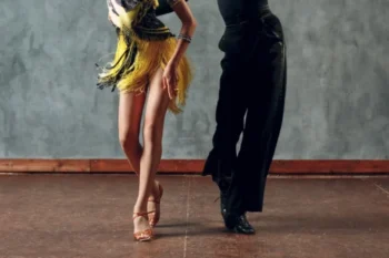 Dva lidé tančí ča-ča. Pár je viditelný od pasu dolů. Žena má na sobě šaty se žlutými třásněmi a hnědé taneční boty. Muž má na sobě černé kalhoty a černé taneční boty. Tančí na dřevěné podlaze před šedou zdí.