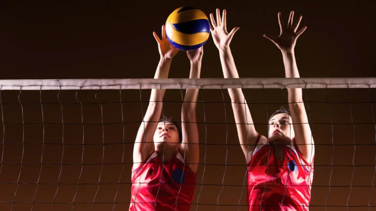 Dva volejbalisté v červených uniformách blokují u sítě během zápasu. Jeden hráč vyskočí výše a dotkne se žlutomodré volejbalové míče rukama nad sítí, zatímco druhý hráč také zvedne ruce do blokovacího postoje.