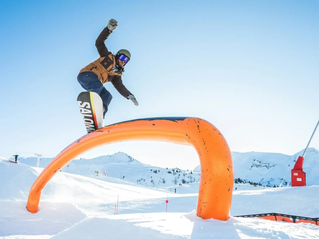 Snowboardista skáče na oranžové hrazdě, skok do výšky se zdviženou rukou, v pozadí zimní hory.